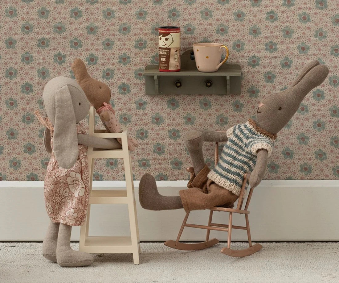 Stuffed rabbit toys in a room with floral wallpaper, chairs and a shelf.