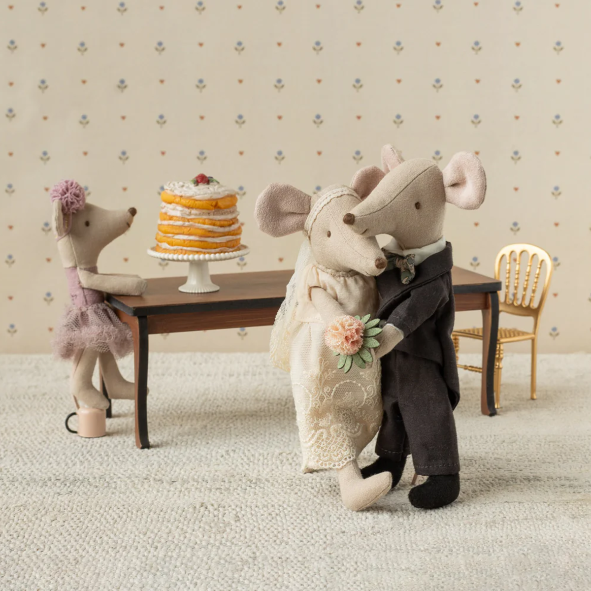 Stuffed mouse couple in wedding attire with a cake on a table against a patterned wall.