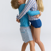 Model wearing red, white and blue  striped boardie swim shorts for him with a blue drawstring and a blue long-sleeve rashguard shirt.  