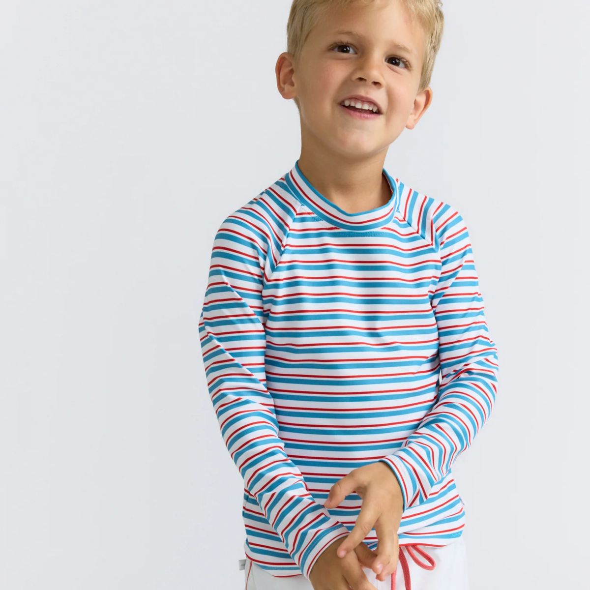 Model wearing striped long-sleeve rashguard top with red, white, and blue colors and white boardie swim shorts with red drawstring