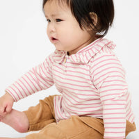 A baby wearing a pink and white striped long-sleeve top with a ruffled collar and brown corduroy pants, sitting against a plain white background.