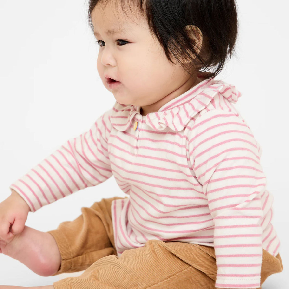 A baby wearing a pink and white striped long-sleeve top with a ruffled collar and brown corduroy pants, sitting against a plain white background.