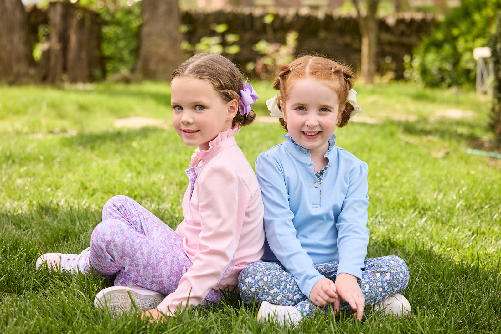 Two young girls sitting back-to-back on a green grass lawn. The girl on the left has light brown hair in a purple bow and wears a pink long-sleeve top with purple patterned leggings.