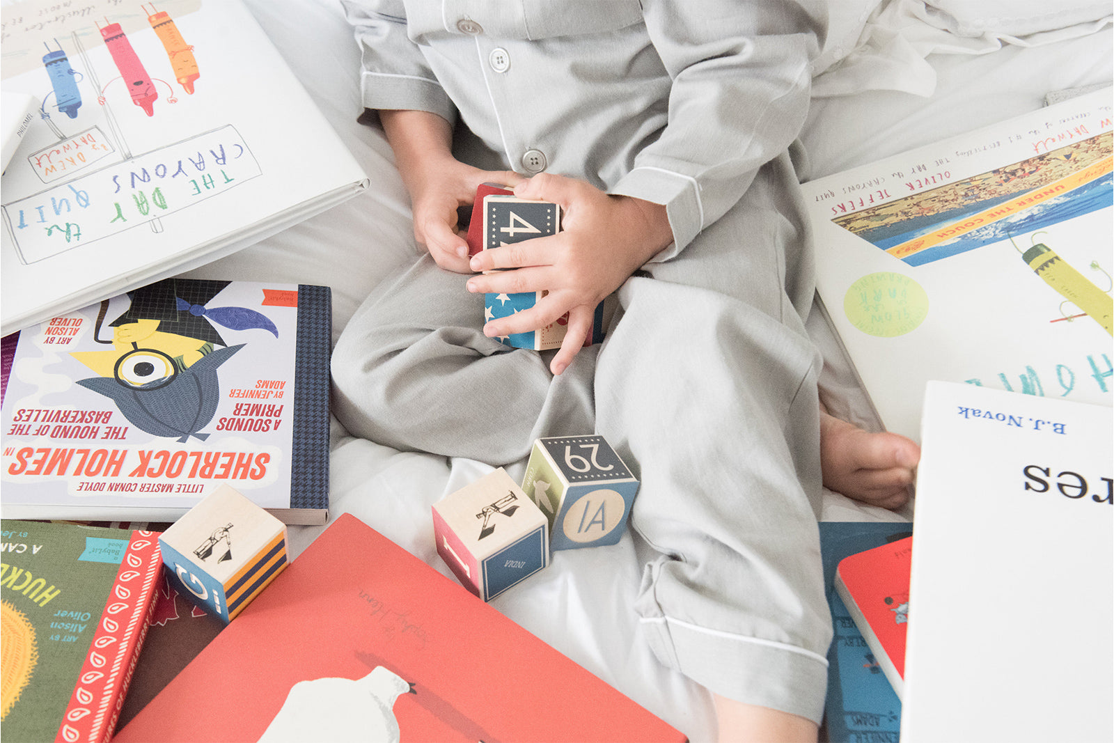 A child wearing light gray pajamas sits on a bed surrounded by books and wooden blocks, holding a colorful toy block.