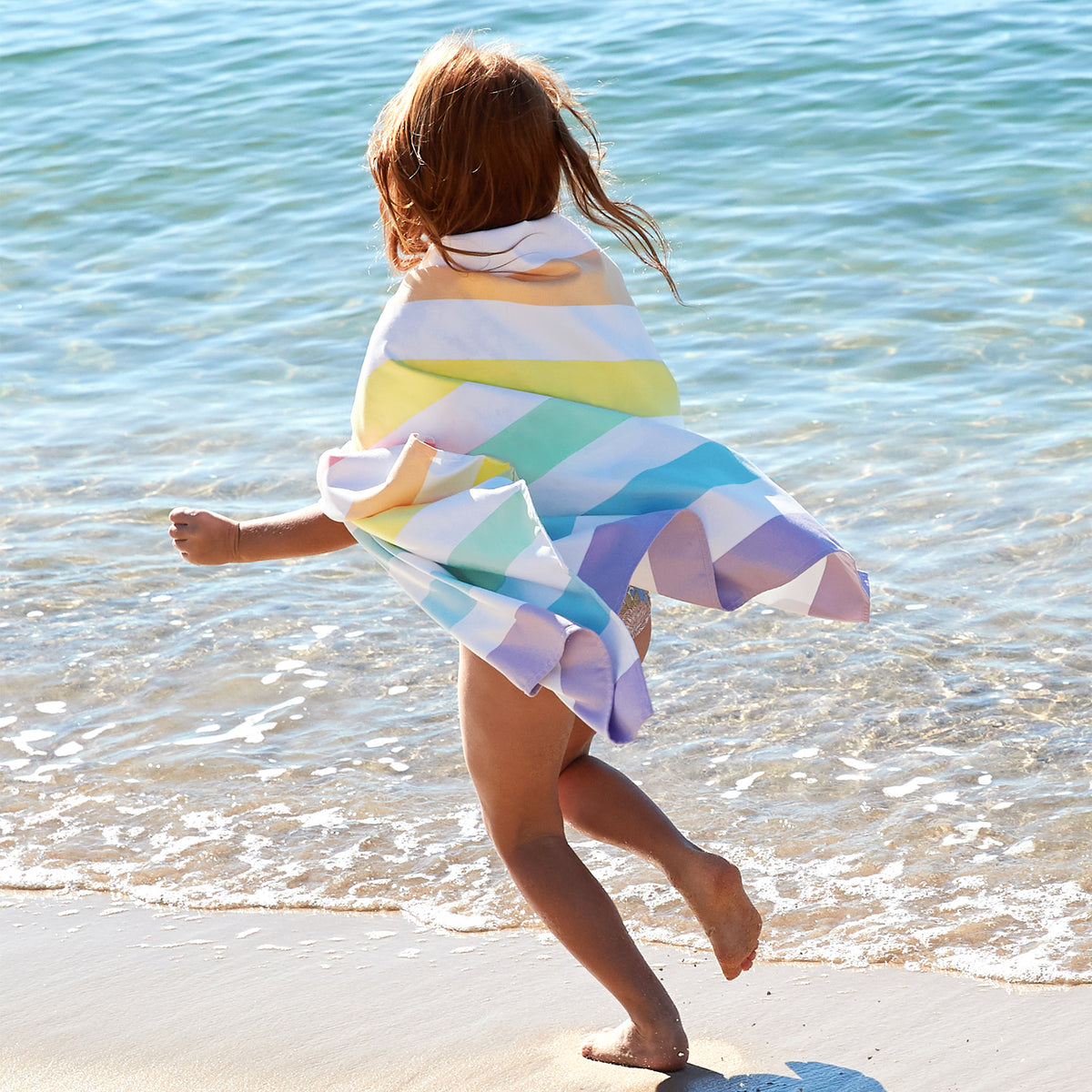 Child running on a beach with a colorful striped towel wrapped around them.