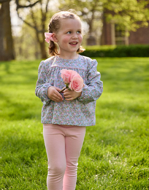 A young girl with a pink hair bow stands on a green lawn holding a small bouquet of pink roses. She is wearing a light blue long-sleeve top with a dense multicolor floral print and light pink leggings.