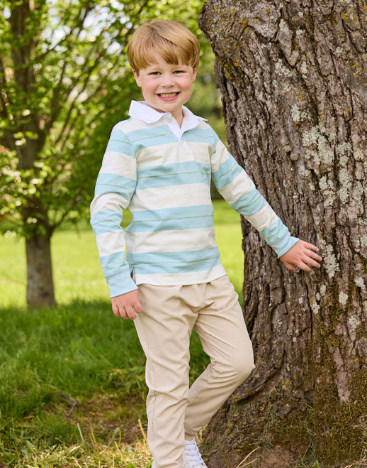 A young boy smiling and posing outdoors next to a large tree trunk, wearing a light blue and white striped long-sleeve rugby shirt with beige trousers.
