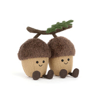 Two smiling acorn plush toys with brown fuzzy caps, tan bodies, dark brown feet, and a small green leaf on a branch above them, photographed on a clean white background.