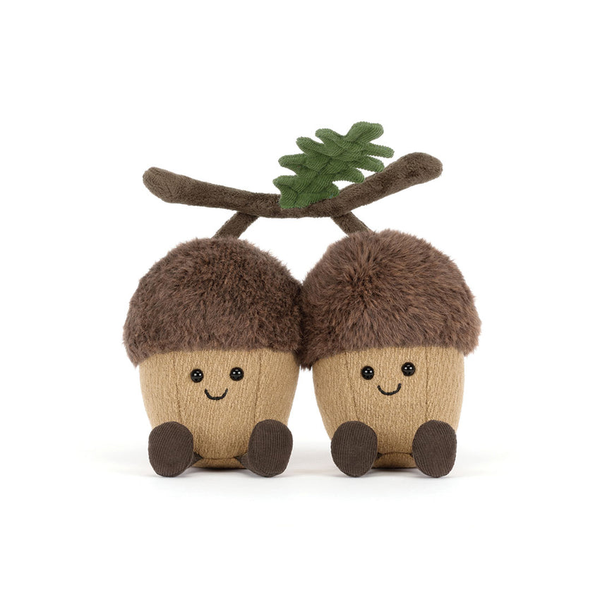 Two plush acorn toys with smiling faces, brown faux-fur caps, tan bodies, dark brown feet, and a small green leaf on a branch above them, photographed on a white background.