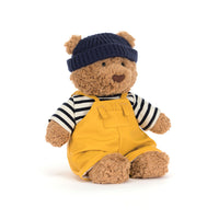 A plush teddy bear wearing a navy blue knit beanie, a black and white striped shirt, and mustard yellow overalls, posed against a clean white background.