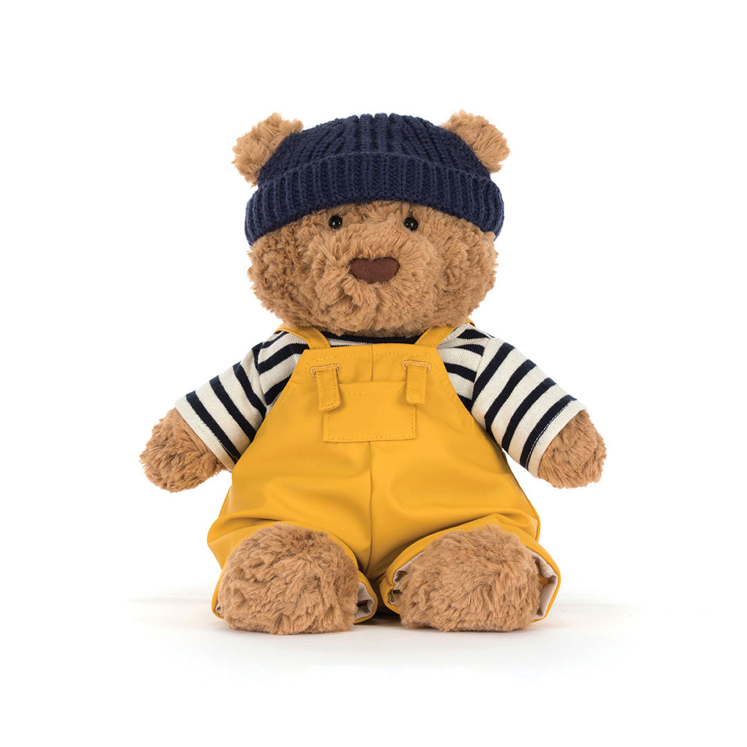 A teddy bear plush toy wearing a navy blue knit beanie, a black and white striped long-sleeve shirt, and mustard yellow overalls, photographed against a clean white background.