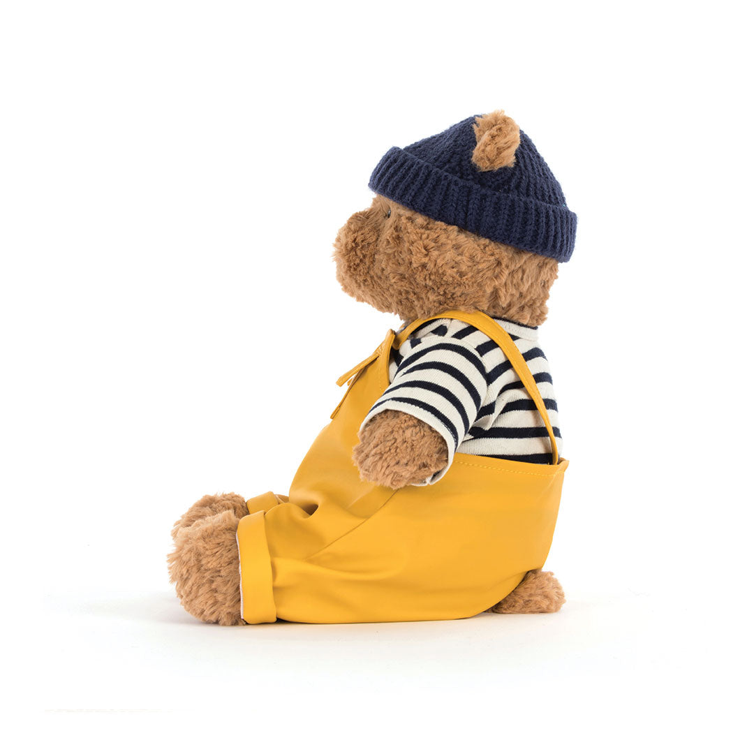 A plush teddy bear doll wearing a navy blue knit beanie, a black and white striped long-sleeve shirt, and mustard yellow overalls, posed against a solid white background.