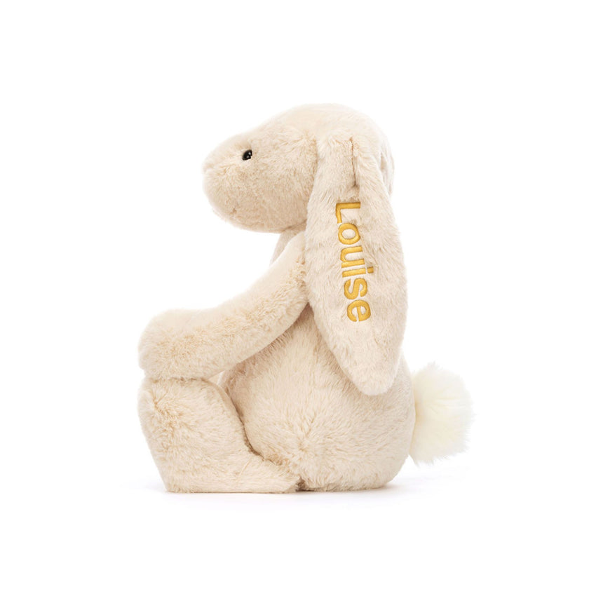 Side view of a cream colored bunny stuffed animal with a personalized ear embroidery on a white background.