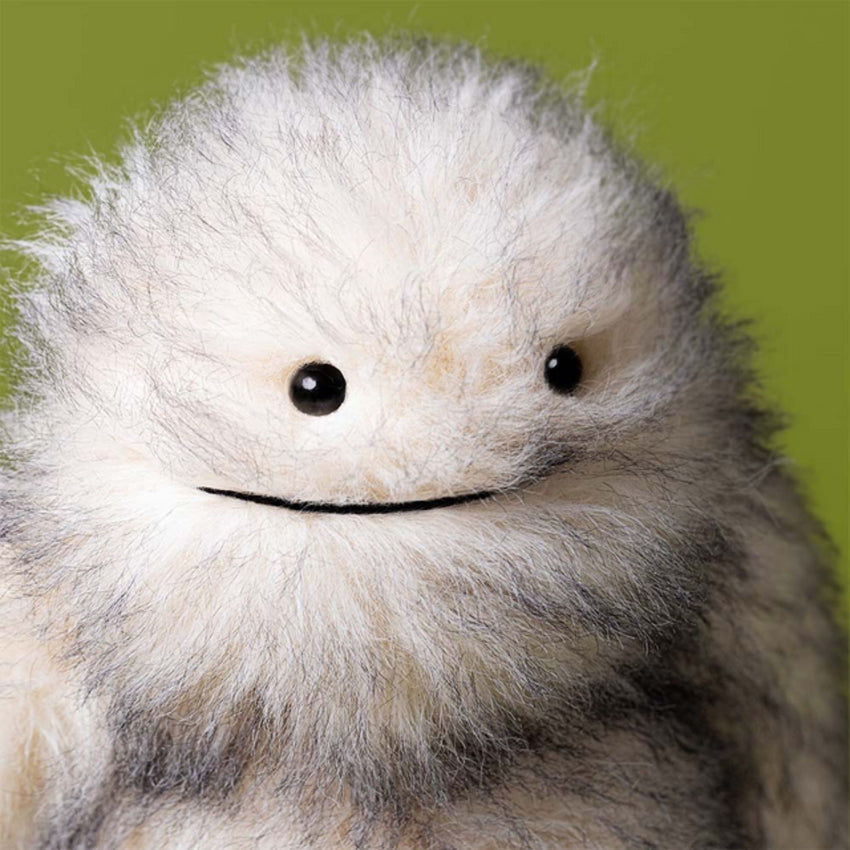 Close up view of a fluffy gray and white furry "yeti" critter plush toy sitting against a plain green background.