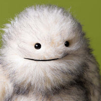 Close up view of a fluffy gray and white furry "yeti" critter plush toy sitting against a plain green background.