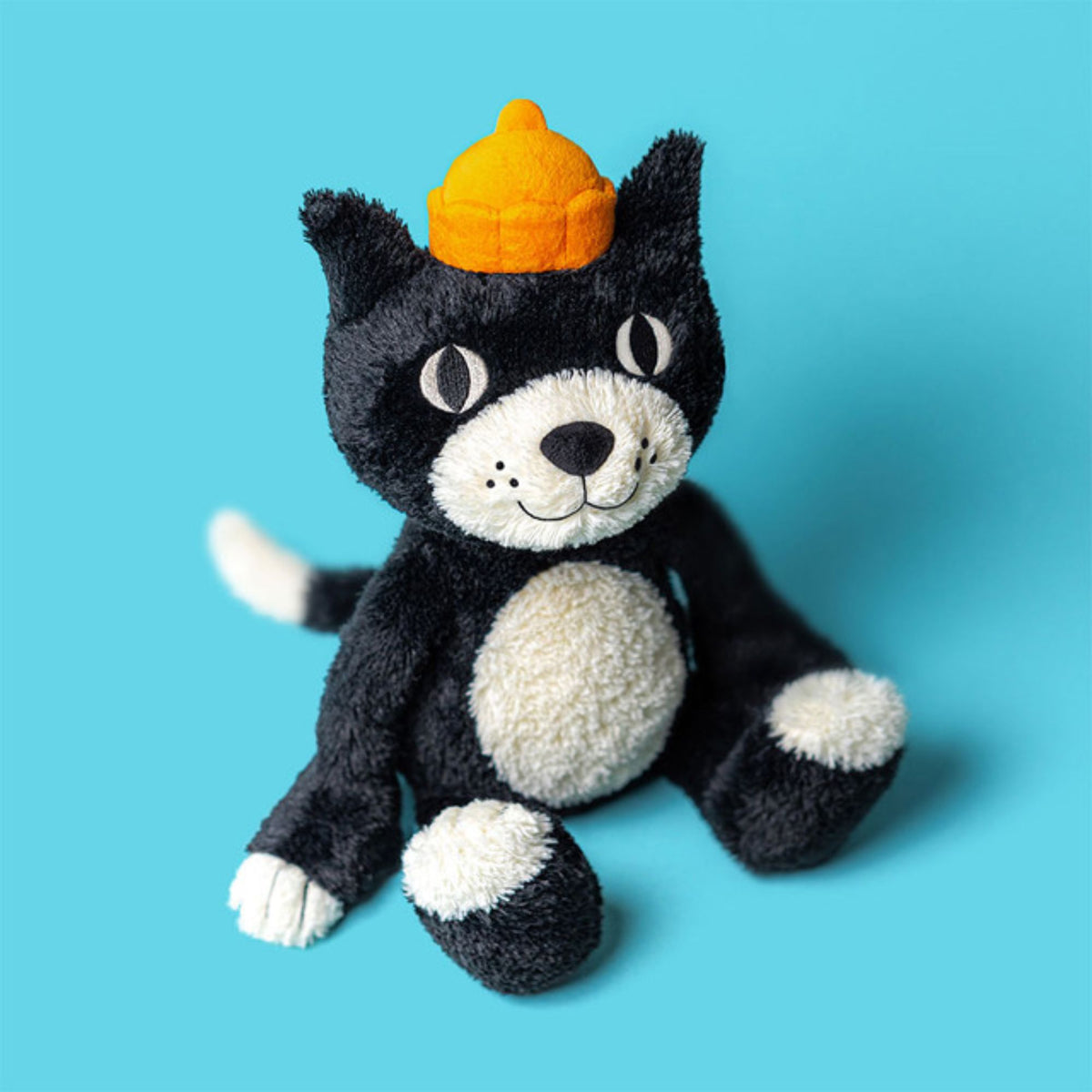 A top view of a black and white plush cat stuffed animal wearing an orange hat on a plain blue background.