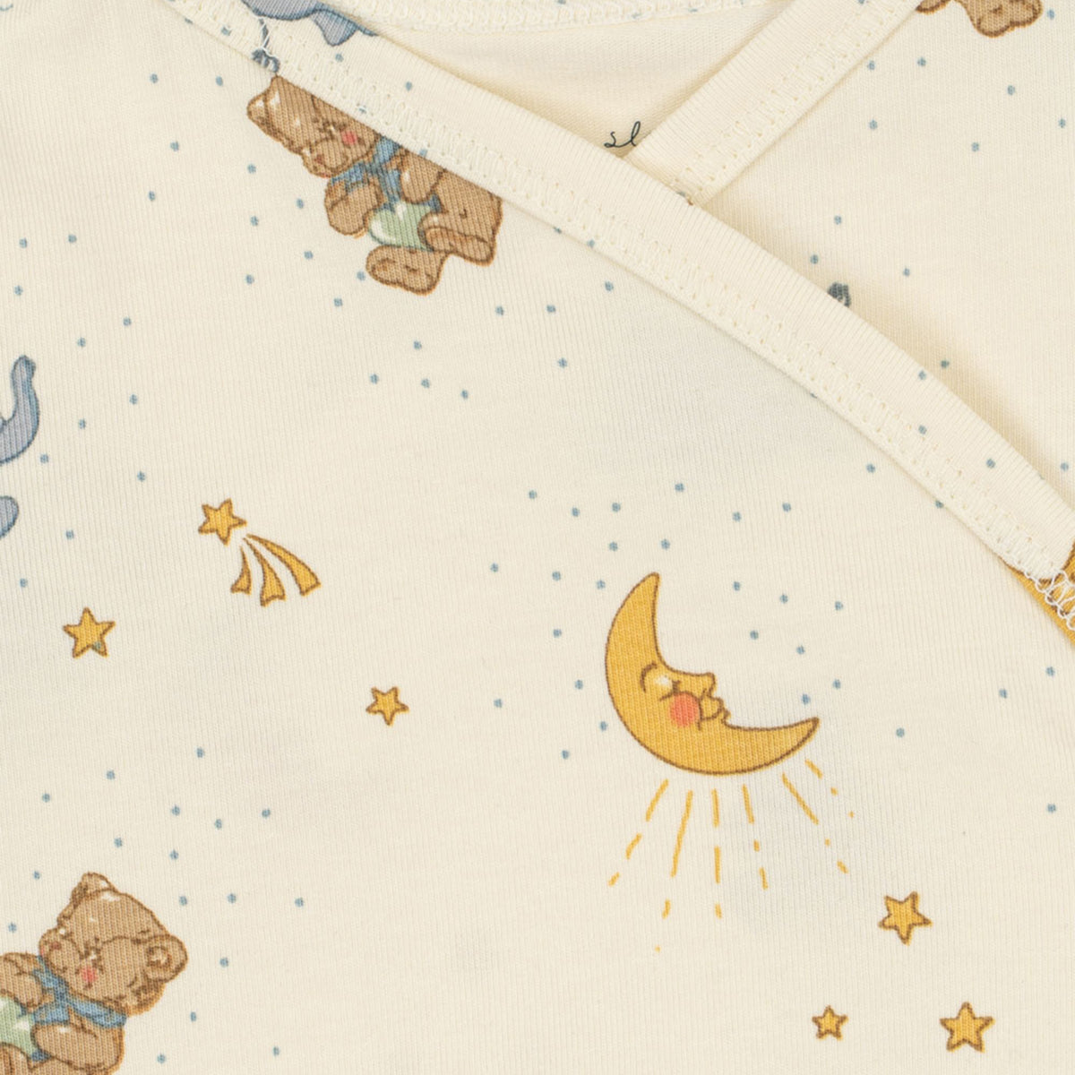 Close-up of a baby body suit in warm off-white, printed with a cute night sky pattern featuring teddy bears, a yellow crescent moon, stars, and small blue dots.