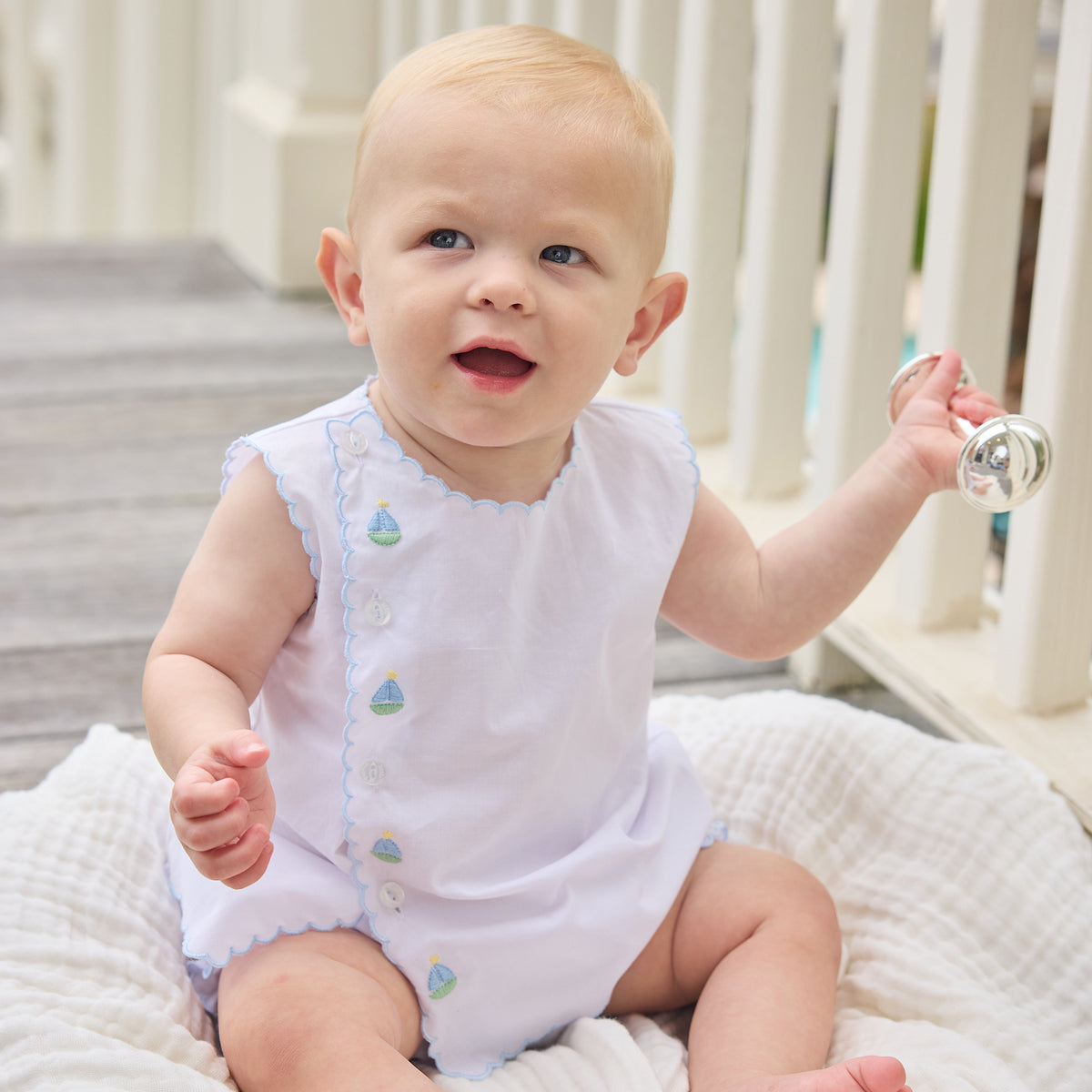Baby sitting on a blanket wearing a white outfit with off-center boat embroidery.