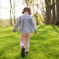 A young girl seen from behind walking on a grassy park path, wearing a light blue floral long-sleeve blouse and pink pants.