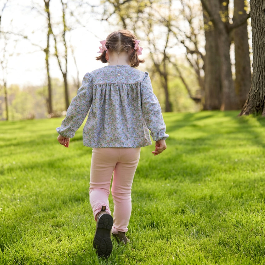 A young girl seen from behind walking on a grassy park path, wearing a light blue floral long-sleeve blouse and pink pants.
