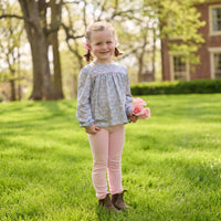 A young girl stands on a sunny green lawn holding a small bouquet of pink roses. She is wearing a light blue and white floral long-sleeve top, pink leggings, and brown boots, with trees and a brick building softly blurred in the background.