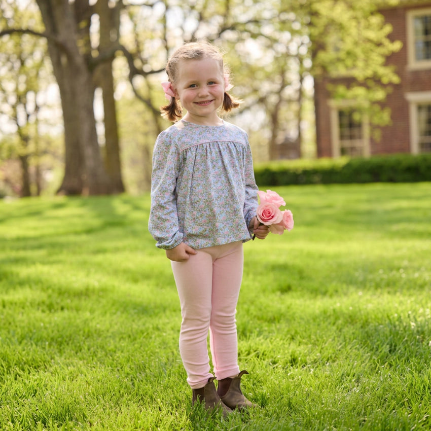 A young girl stands on a sunny green lawn holding a small bouquet of pink roses. She is wearing a light blue and white floral long-sleeve top, pink leggings, and brown boots, with trees and a brick building softly blurred in the background.