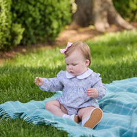 A baby girl sitting on a blue plaid blanket on a green grassy lawn, wearing a light lavender floral long-sleeve romper with a white Peter Pan collar and a pink bow in her hair.