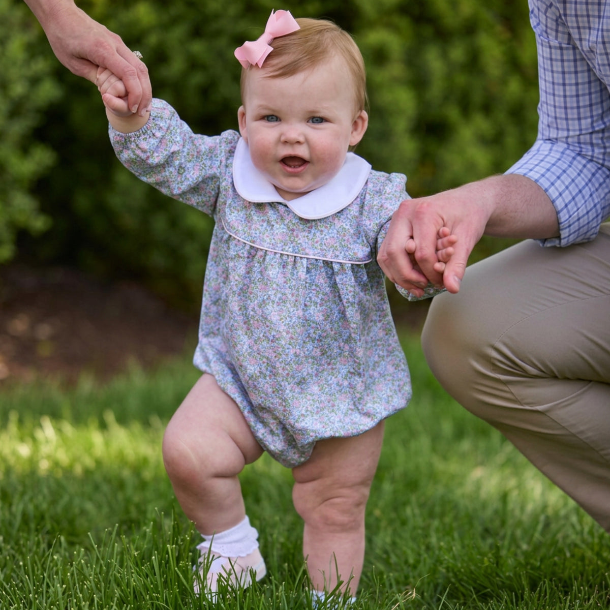 A baby wearing a long-sleeve floral romper with a white Peter Pan collar and a pink bow in her hair, walking outdoors on grass while holding an adult's hand.