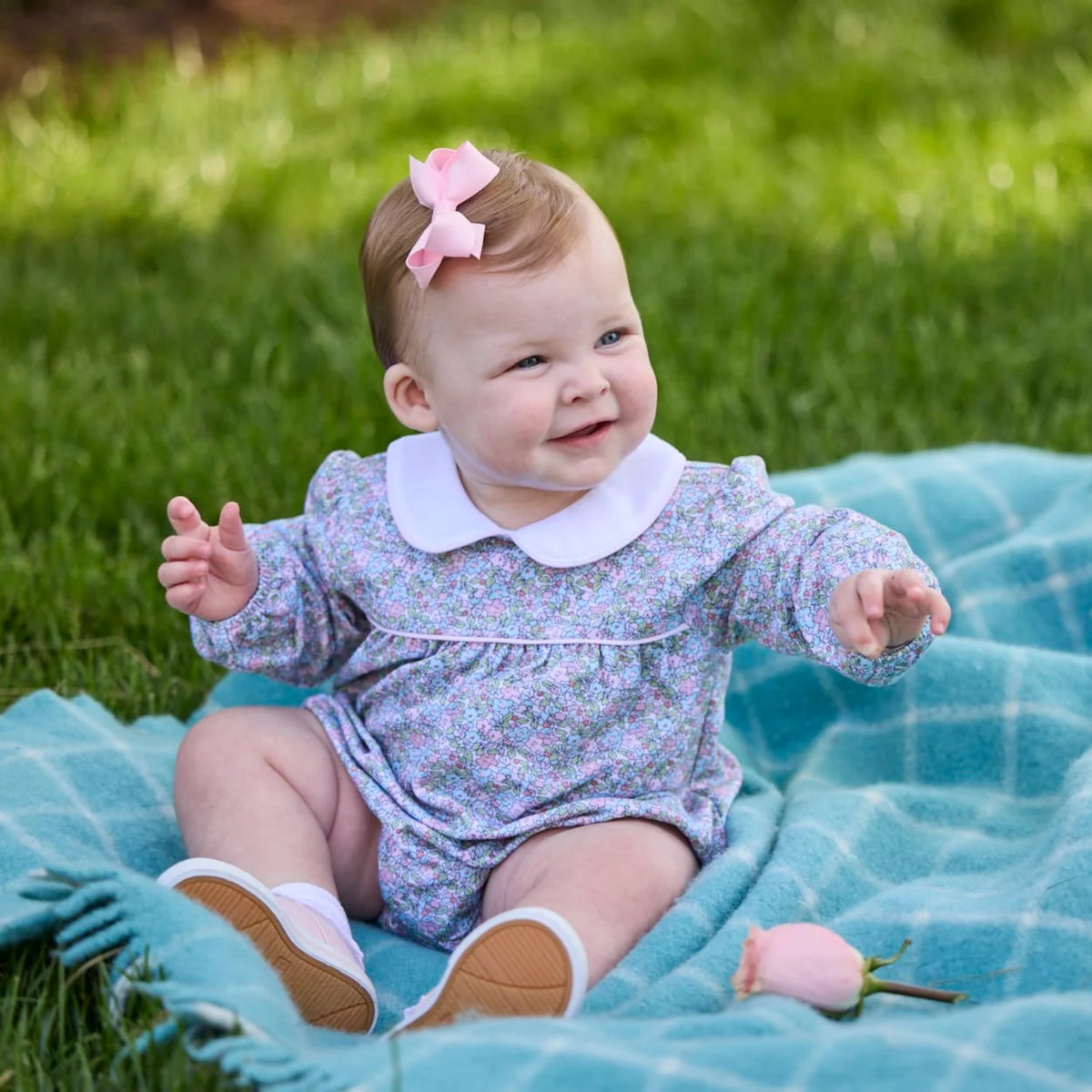 A baby girl sitting on a blue blanket in a grassy park, wearing a lavender floral long-sleeve romper with a white Peter Pan collar, white socks, and a pink bow in her hair.