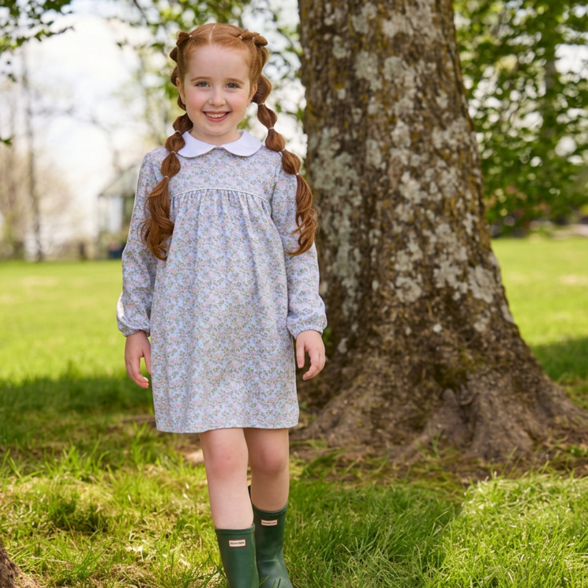 A young girl with long auburn hair in two braids stands outdoors in a grassy park next to a large tree trunk, wearing a light blue and white floral long-sleeve dress with a white collar and dark green rain boots.