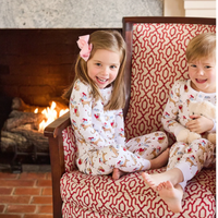 Two children in "I woof you" dog themed pajamas sitting on a patterned chair in front of a fireplace.