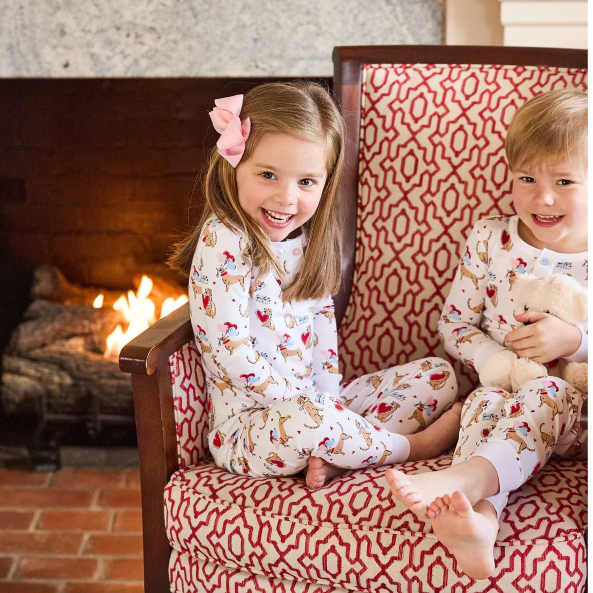 Two children in "I woof you" dog themed pajamas sitting on a patterned chair in front of a fireplace.
