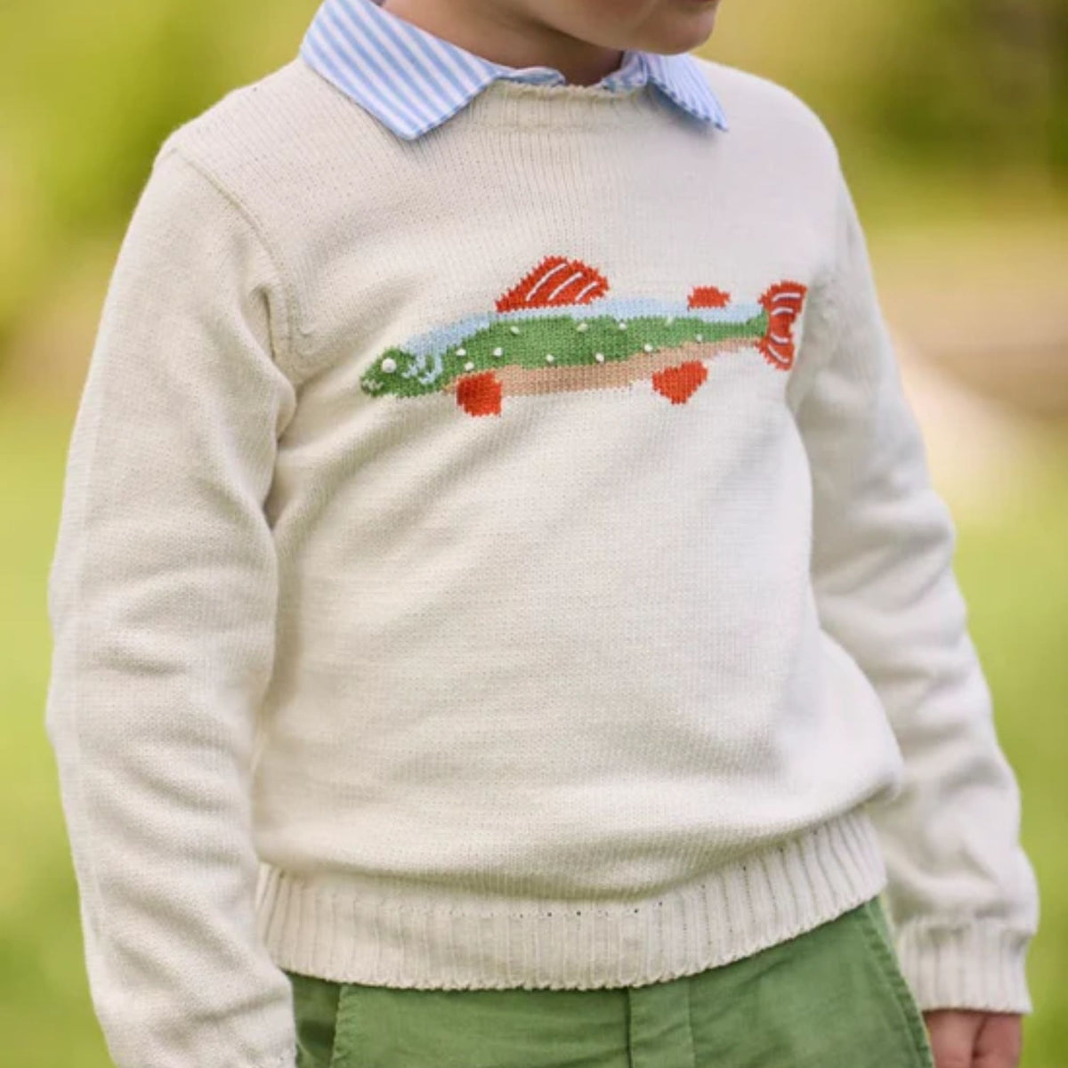A cream colored sweater with an intarsia trout on the chest.  Model wearing green pants in an outdoor setting.