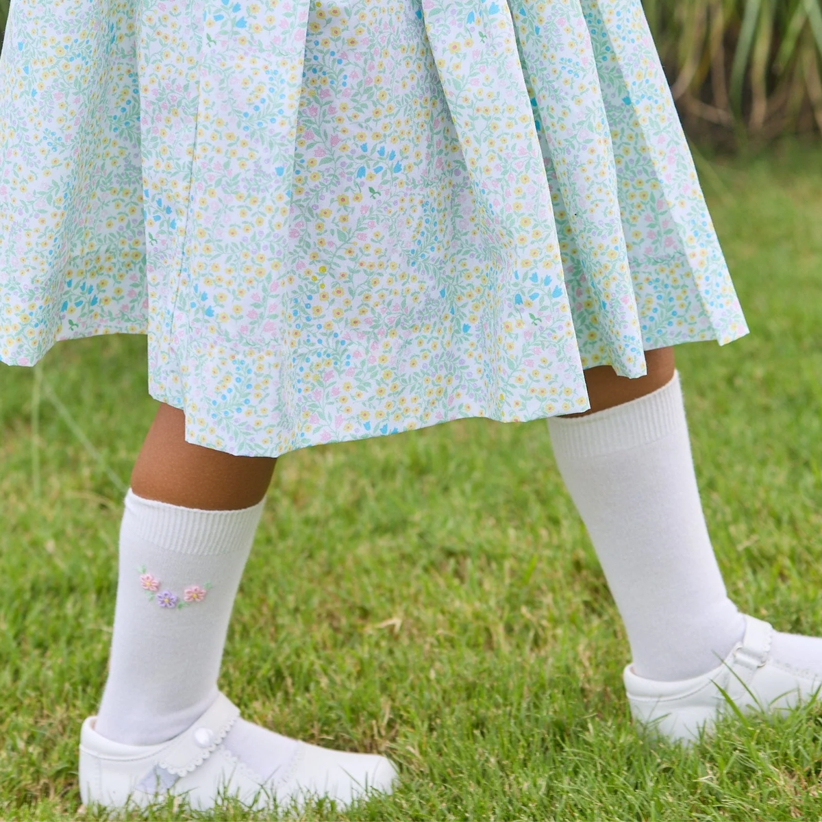Child wearing a floral dress, white socks, and shoes on grass.