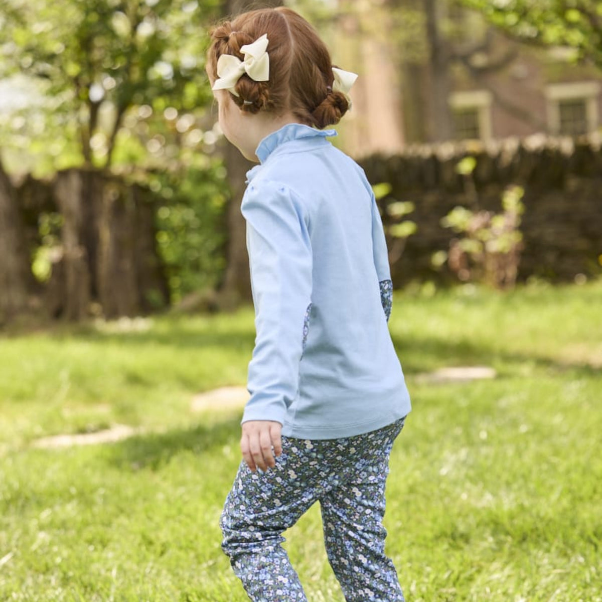 A young child wearing a light blue long-sleeve top and blue floral pants, photographed outdoors on a grassy lawn with trees and a fence in the background.