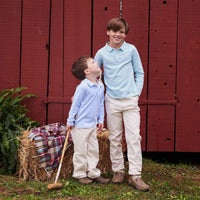 Two boys wearing matching light blue striped polo shirts and beige pants standing in front of a red wooden barn door, with a hay bale and plaid blanket nearby.