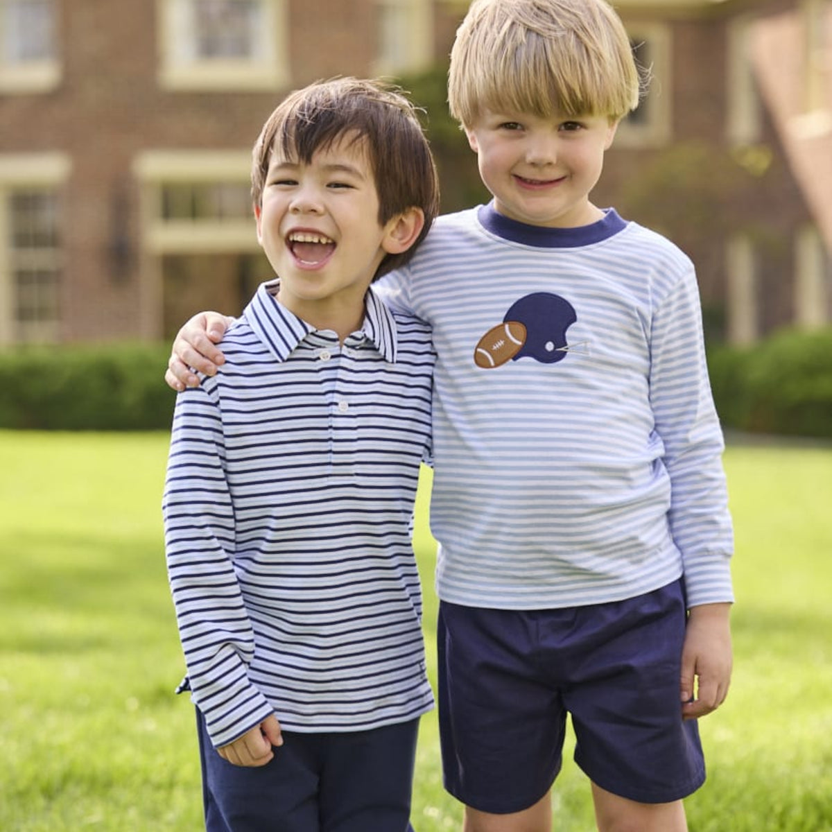 Two young boys standing outdoors on a grassy lawn in front of a brick building. The boy on the left wears a navy and white striped long-sleeve polo shirt with navy shorts, and the boy on the right wears a light blue and white striped long-sleeve shirt with a navy football helmet and brown football graphic on the chest, paired with navy shorts.