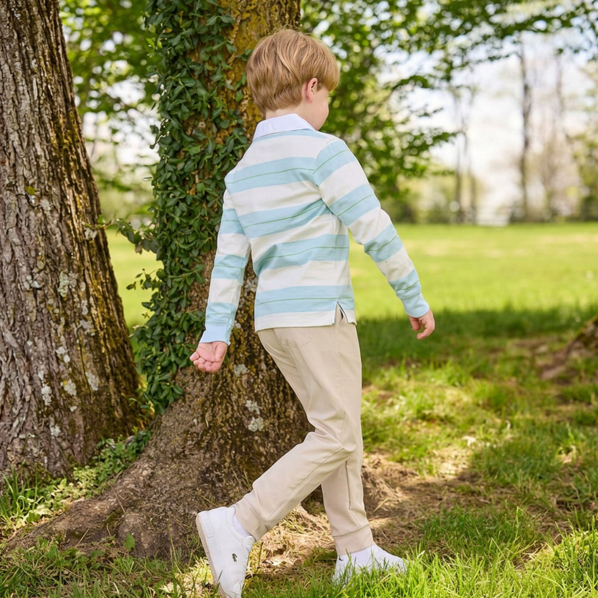 A young boy with blond hair is seen from behind, walking outdoors next to a large tree trunk in a grassy park. He is wearing a light blue and white striped long-sleeve polo shirt, beige trousers, and white sneakers.