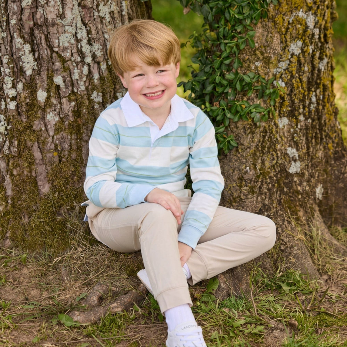 Boy sitting by tree wearing rugby style shirt and khaki pants.