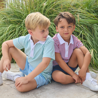 Two young boys wearing striped polo shirts and shorts sitting on a stone path with green grass in the background.