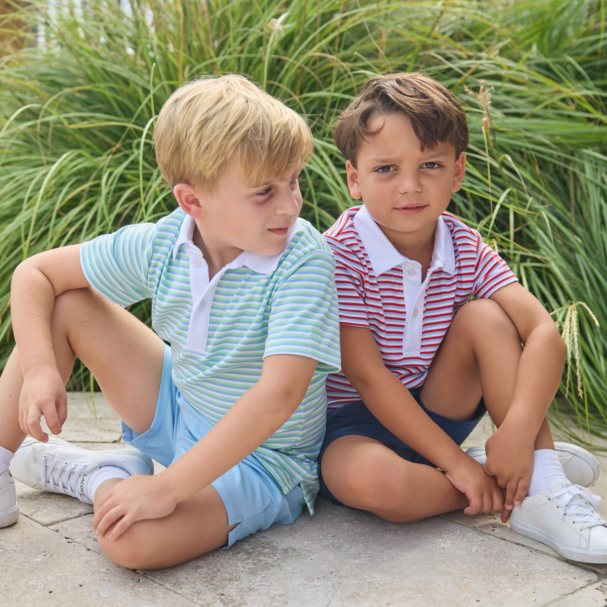 Two young boys wearing striped polo shirts and shorts sitting on a stone path with green grass in the background.