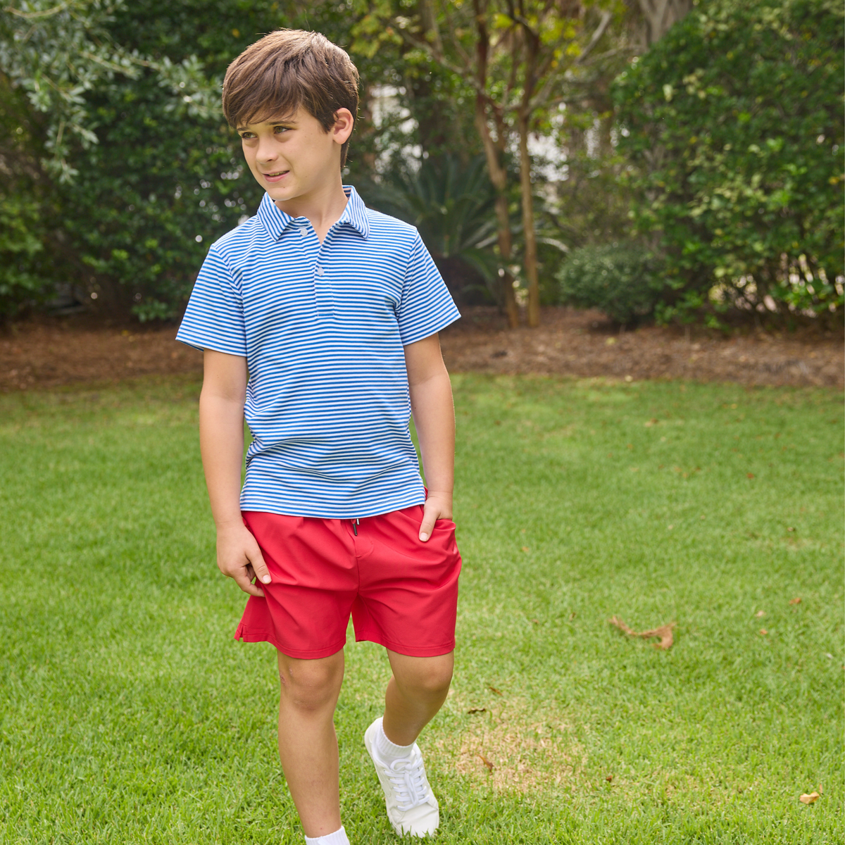 Young boy wearing a blue and white striped polo shirt and red shorts standing on grass.