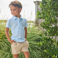 Young boy wearing a light blue striped polo shirt and beige shorts standing outdoors with greenery around.