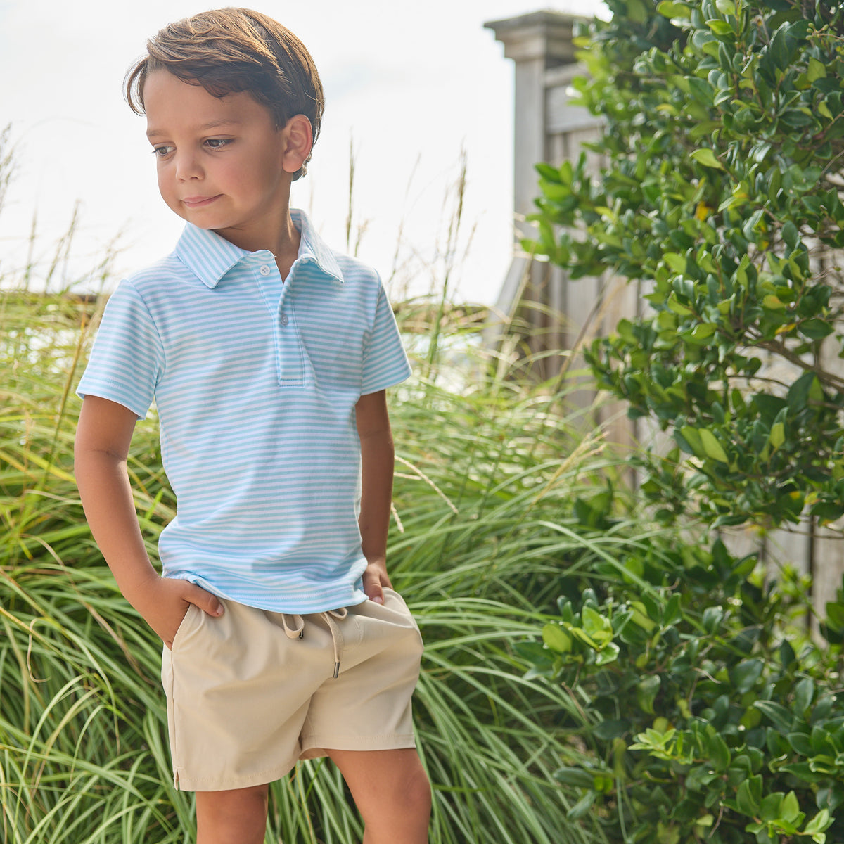 Young boy wearing a light blue striped polo shirt and beige shorts standing outdoors with greenery around.