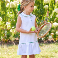 Young girl in a white tennis dress with green trim holding a tennis racket and ball in a garden.