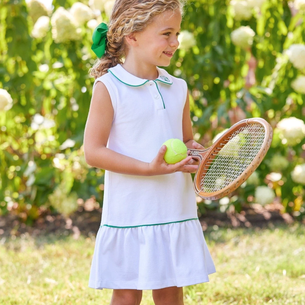Young girl in a white tennis dress with green trim holding a tennis racket and ball in a garden.