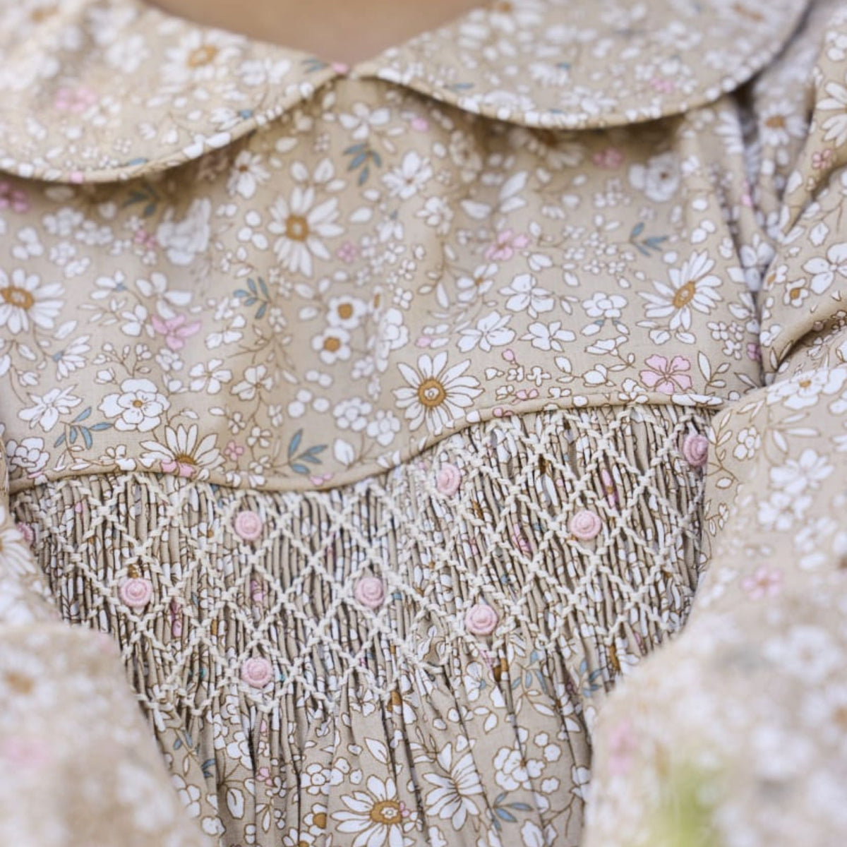 Close-up of a beige floral dress with a Peter Pan collar, delicate white and pink daisy print, and a smocked bodice with embroidered details.