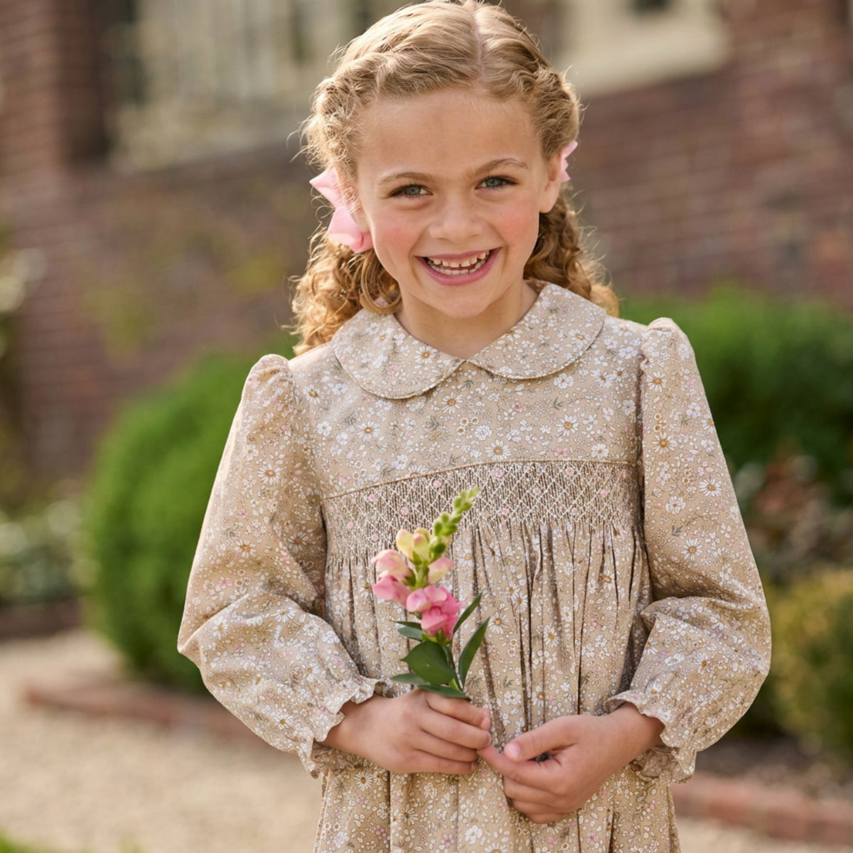 A young girl with curly blonde hair smiles while wearing a beige and white floral-patterned long-sleeve dress with a smocked chest panel. She is holding a small pink flower sprig in an outdoor garden setting.