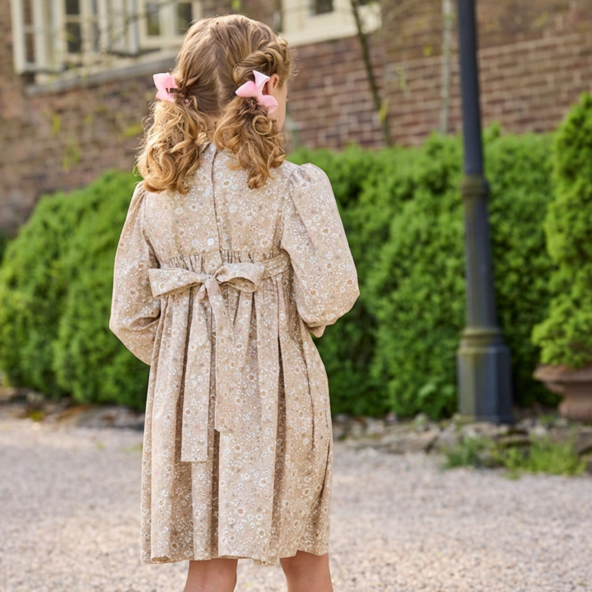 A young girl with curly blonde hair tied back with pink bows is shown from behind, wearing a beige long-sleeve dress with a small white floral pattern and a matching waist tie, standing outdoors in front of green hedges and a brick wall.