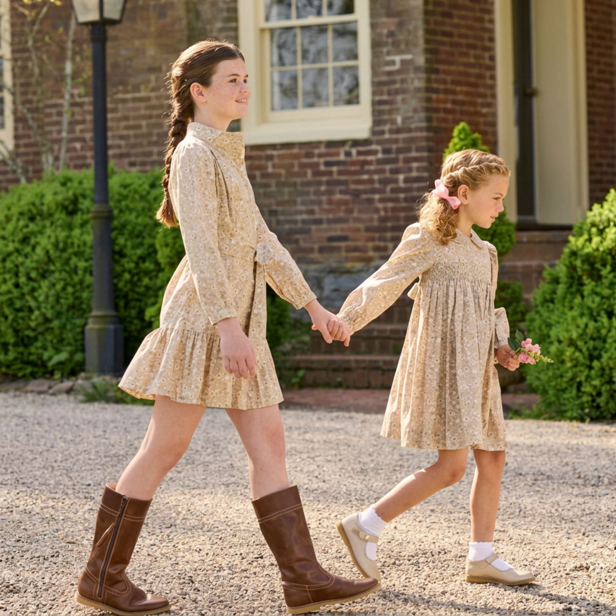 Two young girls walking hand in hand on a gravel path in front of a brick house. Both are wearing matching beige floral long-sleeve dresses and brown boots.